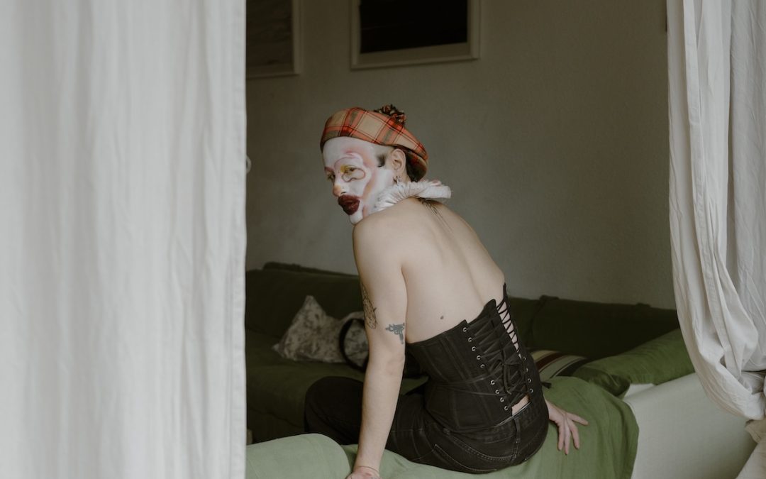 A person in drag makeup is sitting on a bed, their back turned to the camera, looking back over their shoulder