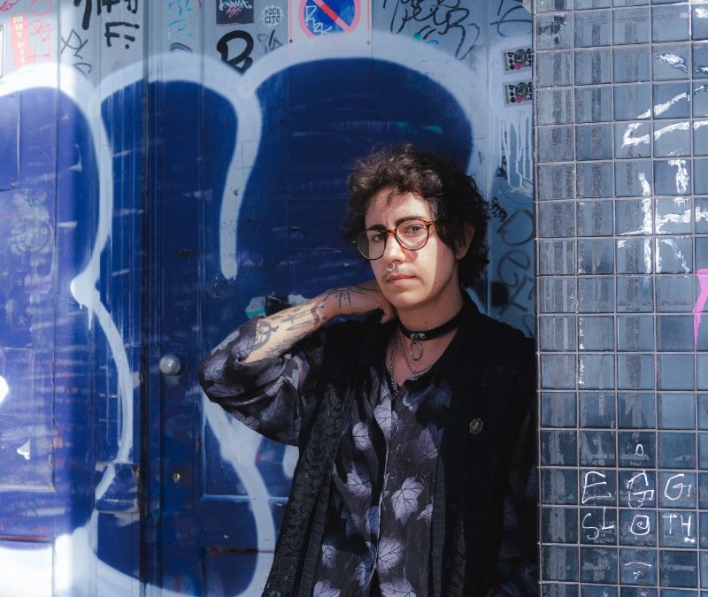 A person in dark-tone clothes posing for a photo in front of a graffiti-inscribed wall