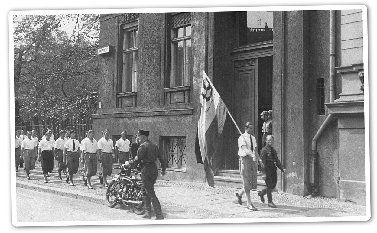 A black and white photo of a group of young men in white shirts, with a flag carried in front of them